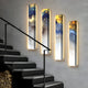 A staircase features four vertically mounted Chroma Gold Radiant Veins LED Wall Art Canvas pieces by Giant Sculptures, glowing luxuriously. The abstract blue, yellow, and white art resembles clouds. An elegant ascent is marked by a black handrail, with logs resting below.