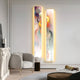 A modern living room with a light gray armchair, a small round table, and an ornate mantle. The wall features two tall Chroma Mist Fog Details LED Wall Art Canvas pieces by Giant Sculptures, casting a warm glow with their premium soft backlighting and adding color to the contemporary interior.