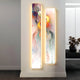 The Chroma Mist Fog Details LED Wall Art Canvas by Giant Sculptures, featuring swirling reds, oranges, and purples, adorns a white wall. The panels are highlighted by soft lights. A black side table with dried flowers enhances the contemporary interior ambiance.