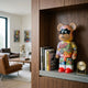 Lucky Paws Bear Sculpture, 50cm, resin bear with masked face and graffiti-style "Hello" artwork, displayed on a wooden shelf beside books in a living room.