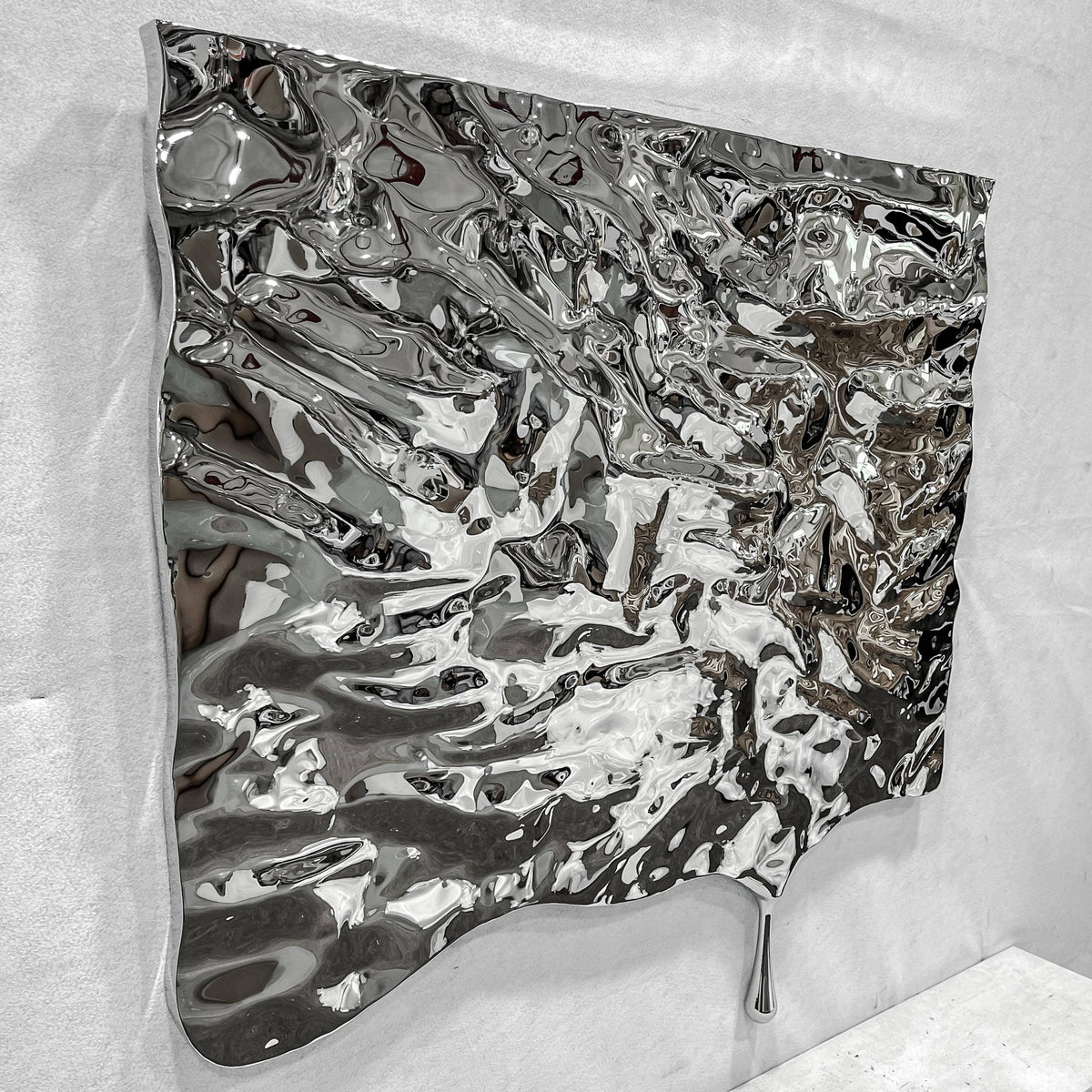 Lumineris Silver Melted Wave Metal 3D Wall Art in mirror-polished stainless steel, shown at an angle with a rippled, liquid-like surface and elongated drip, mounted on a plain white wall.