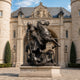 Giant Sculptures' 260cm bronze knight in plate armour lances a coiled dragon beside a rearing horse, dark patina, on a stone plinth before a Gothic building.
