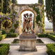 Giant Sculptures Large Classical Three Graces Bronze Sculpture 220cm on a dark marble plinth in a formal garden with archway, wisteria, and cypress trees.