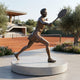 Large Contemporary Female Tennis Player Bronze Sculpture by Giant Sculptures, 210cm, mid-swing lunge with racket raised, on a round pale stone plinth beside a clay court.