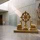 Large Traditional Recumbent Deer and Dharma Wheel Bronze Sculpture by Giant Sculptures in polished gold, displayed in a concrete-walled gallery interior.