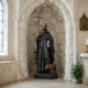 Large Traditional Robed Saint Bronze Statue by Giant Sculptures displayed in a stone archway niche, holding a cross staff and book with a reddish-brown pig at his feet.