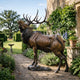 Life-Size Bugling Bull Elk Bronze Sculpture by Giant Sculptures on a stone plinth, head raised mid-call, displayed on a garden path beside lavender.