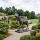 Life-Size Bull Moose Bronze Sculpture by Giant Sculptures with dark patina and wide palmate antlers, on a stone base in a gravel garden path beside roses and wisteria.
