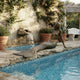 Life-Size Classical Reclining Mermaid Bronze Sculpture by Giant Sculptures resting on stone poolside with verdigris tail, beside a waterfall and bougainvillea.