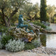 Life-Size Classical Seated Mermaid on Rock Bronze Sculpture by Giant Sculptures with verdigris patina, seated on a mossy boulder beside a garden pond with olive trees.