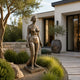 Life-Size Classical Standing Nude Female Bronze Sculpture by Giant Sculptures with dark brown patina, displayed on a gravel bed beside an olive tree at a modern home entrance.