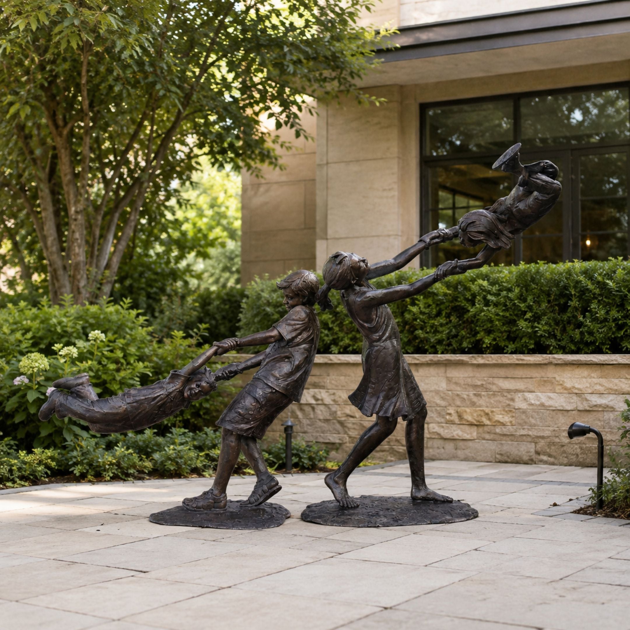 Life-Size Contemporary Children Playing Bronze Sculpture, 200cm, showing two figures swinging a third child through the air, set on a stone patio near greenery.