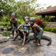 Life-Size Contemporary Children and Crocodile Bronze Sculpture, 150cm, showing four children playing on a bronze crocodile beside a garden pond.