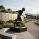 Life-Size Contemporary Male Runner Bronze Sculpture by Giant Sculptures on a black plinth beside a landscaped pathway, with a stadium visible in the background.