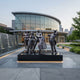 Life-Size Contemporary Musicians and Singing Children Bronze Sculpture by Giant Sculptures, 240cm ensemble with double bass, violin, flute and children, on a black plinth outside a glass concert hall.