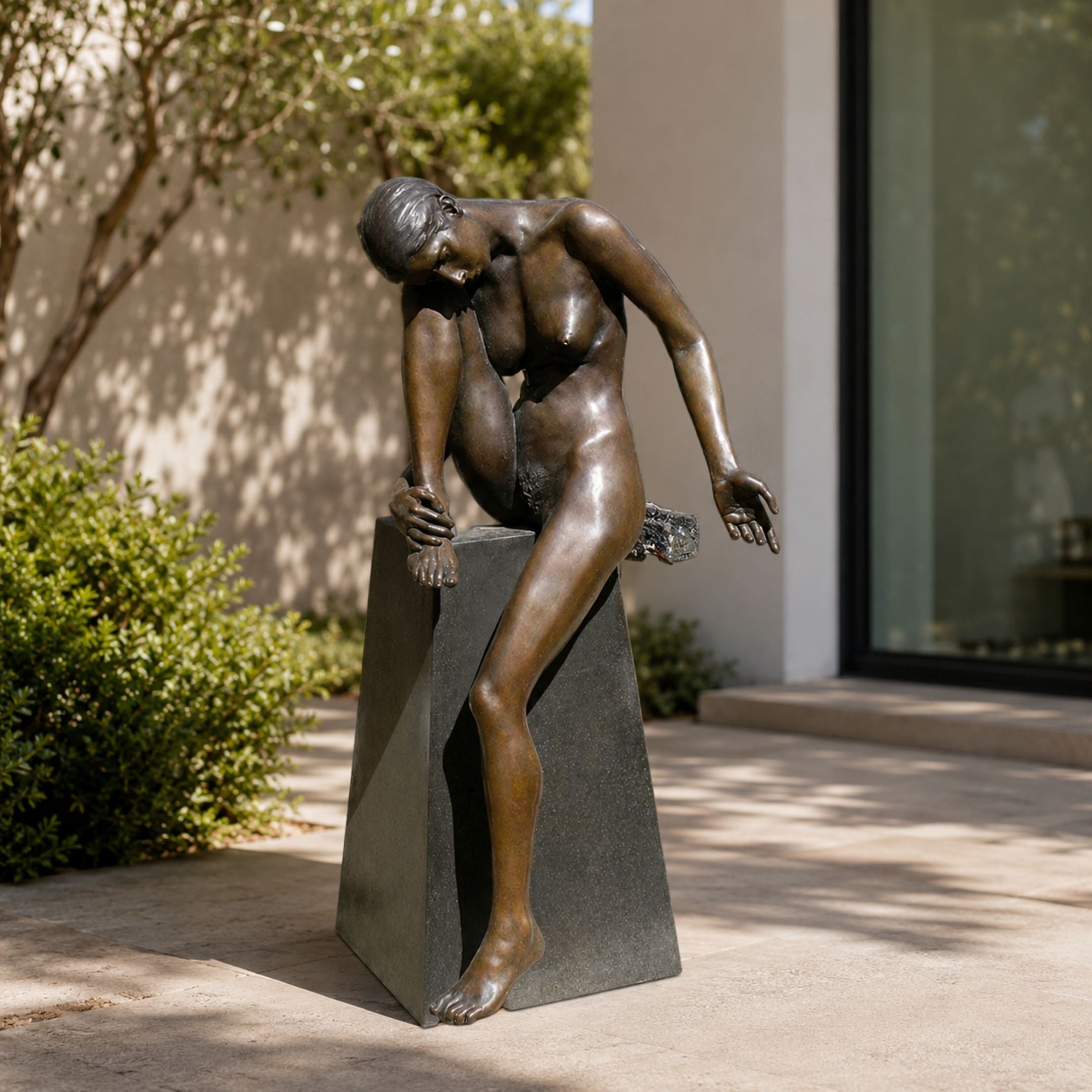 Life-Size Contemporary Seated Female Figure Bronze Sculpture by Giant Sculptures on a dark granite trapezoidal plinth in a sunlit courtyard beside greenery.