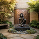 Life-Size Contemporary Seated Nude Female Bronze Sculpture by Giant Sculptures on a black plinth in a courtyard with water wall, ferns, and succulents.