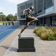 Life-Size Female Sprinter Bronze Sculpture by Giant Sculptures on a black plinth beside a blue running track, mid-stride with warm brown patina and verdigris accents.