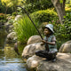 Life-Size Traditional Boy Fishing Bronze Sculpture, 130cm, by Giant Sculptures, showing a crouched boy in a hat holding a fishing rod with a caught fish, set on rocks beside a garden pond.