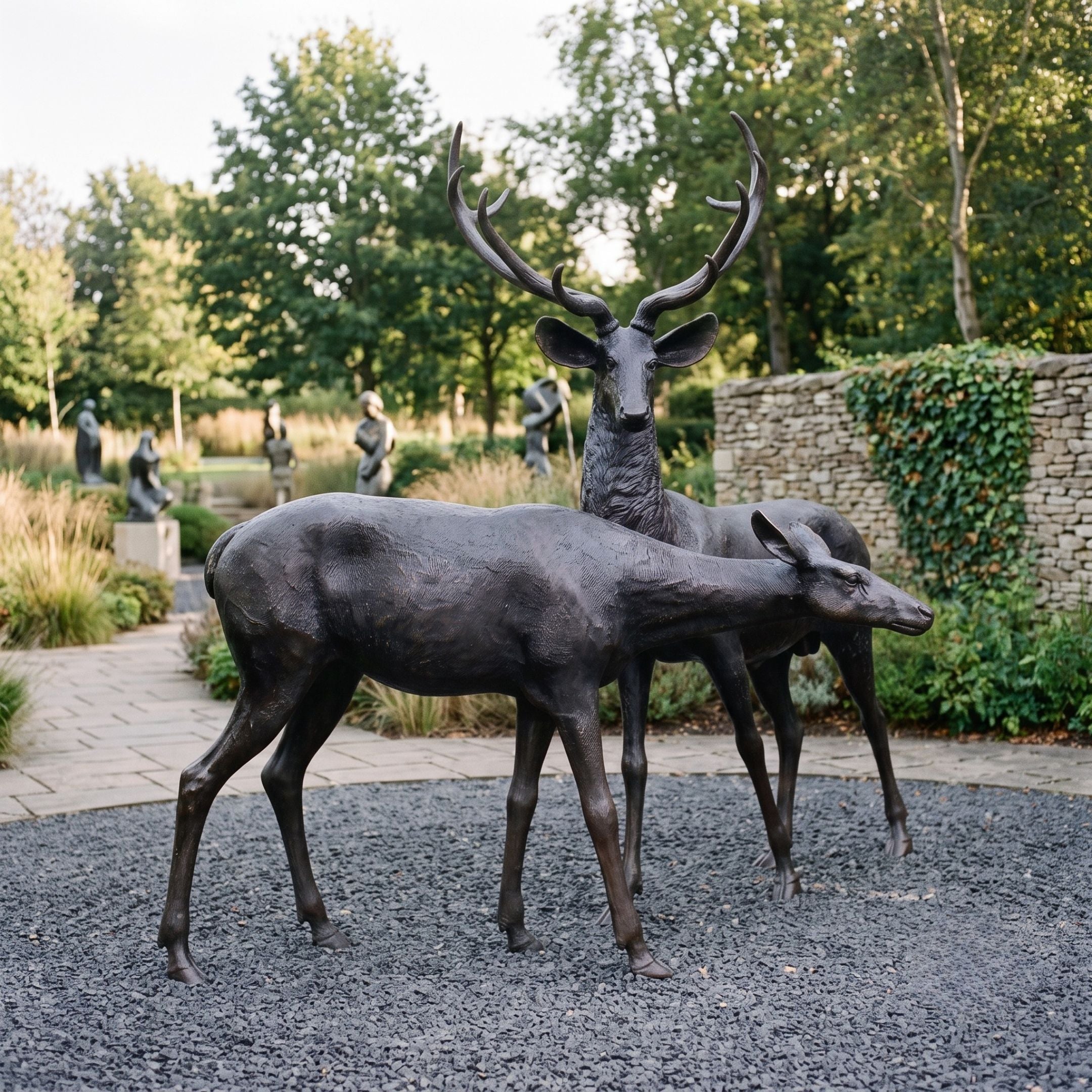 Life-Size Traditional Stag and Doe Deer Bronze Sculpture by Giant Sculptures in dark patina, shown on gravel in a sculpture garden with stone wall behind.