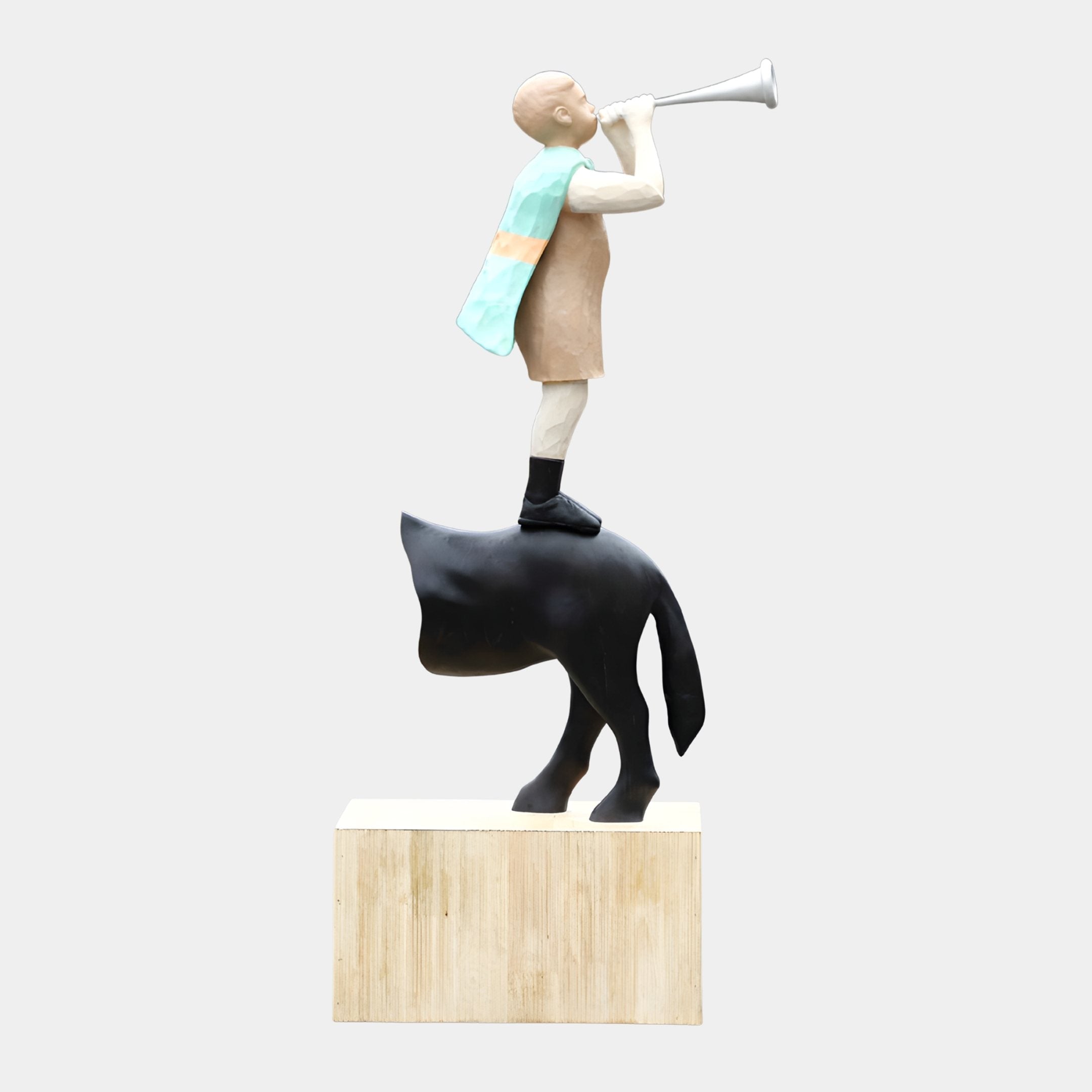 Boy on Buffalo Trumpet Player Wood Sculpture by Giant Sculptures, 160cm hand-carved figure in tan tunic and mint cape raising a silver horn atop a matte black buffalo on a raw wood plinth.