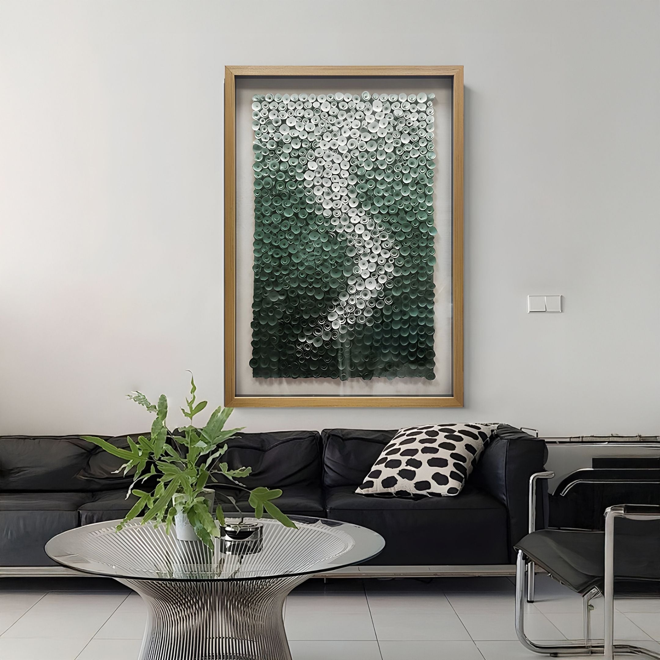 A modern living room with a black sofa, a black-and-white spotted pillow, a glass coffee table topped with a green leafy plant, and the Spiravale Green Gradient Floral Curl 3D Wall Art by Giant Sculptures on a light gray wall.