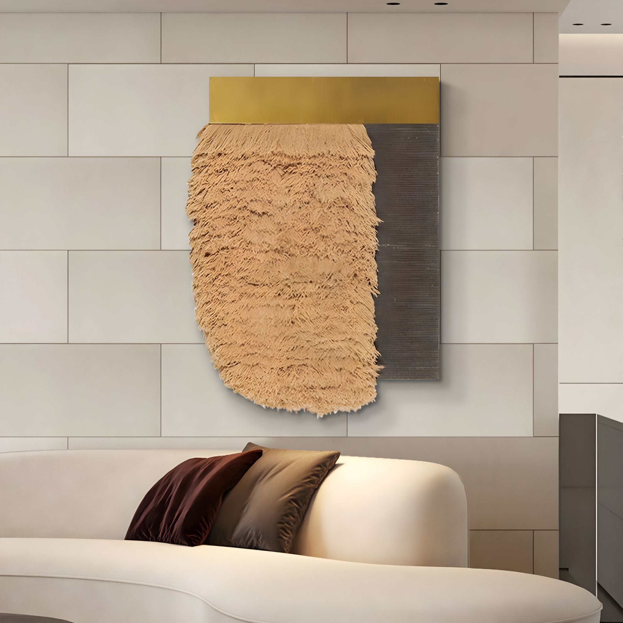 The Halvoria Grey & Sand Fibre Panel 3D Wall Art by Giant Sculptures hangs on a tiled wall above a white curved sofa with three pillows, showcasing tan textured fringe, a metallic gold top, and a dark rectangular panel.