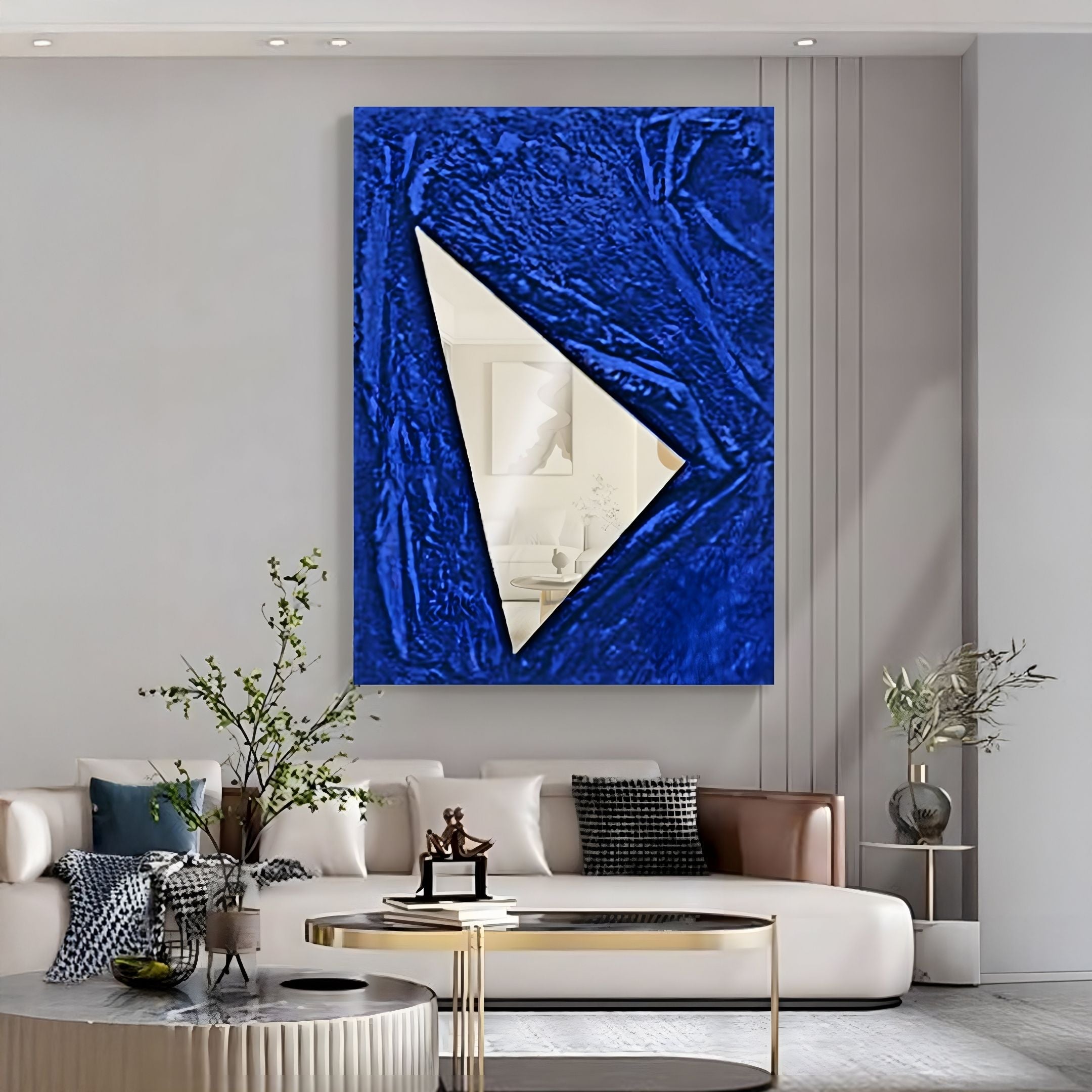 A modern living room with a white sofa, glass coffee table, and plants. Above the sofa is the Arclunor Cobalt Blue Triangle Relief Mirror 3D Wall Art by Giant Sculptures, reflecting part of the space.