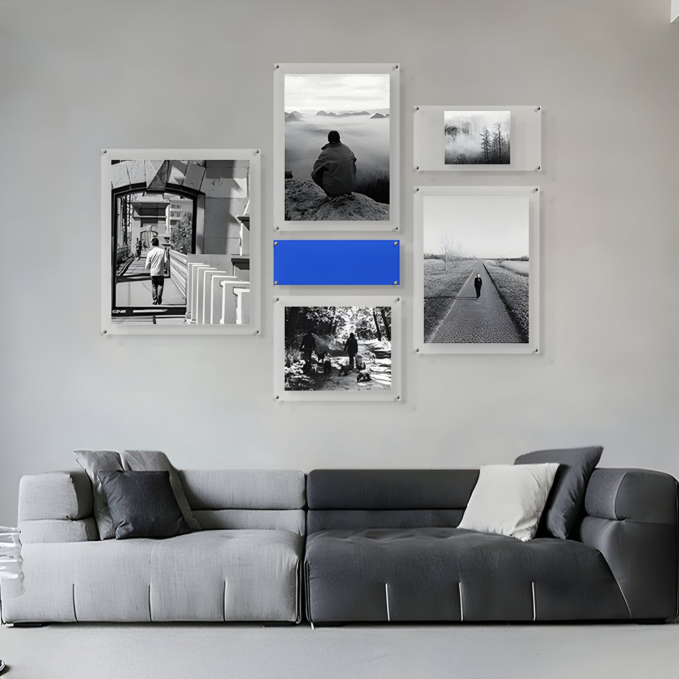 A modern gray sofa faces a light gray wall decorated with the Giant Sculptures Framegrid Blue Mixed-Size Gallery 6-Set Acrylic Wall Art, featuring black-and-white photos and a bold blue rectangle for a contemporary look.