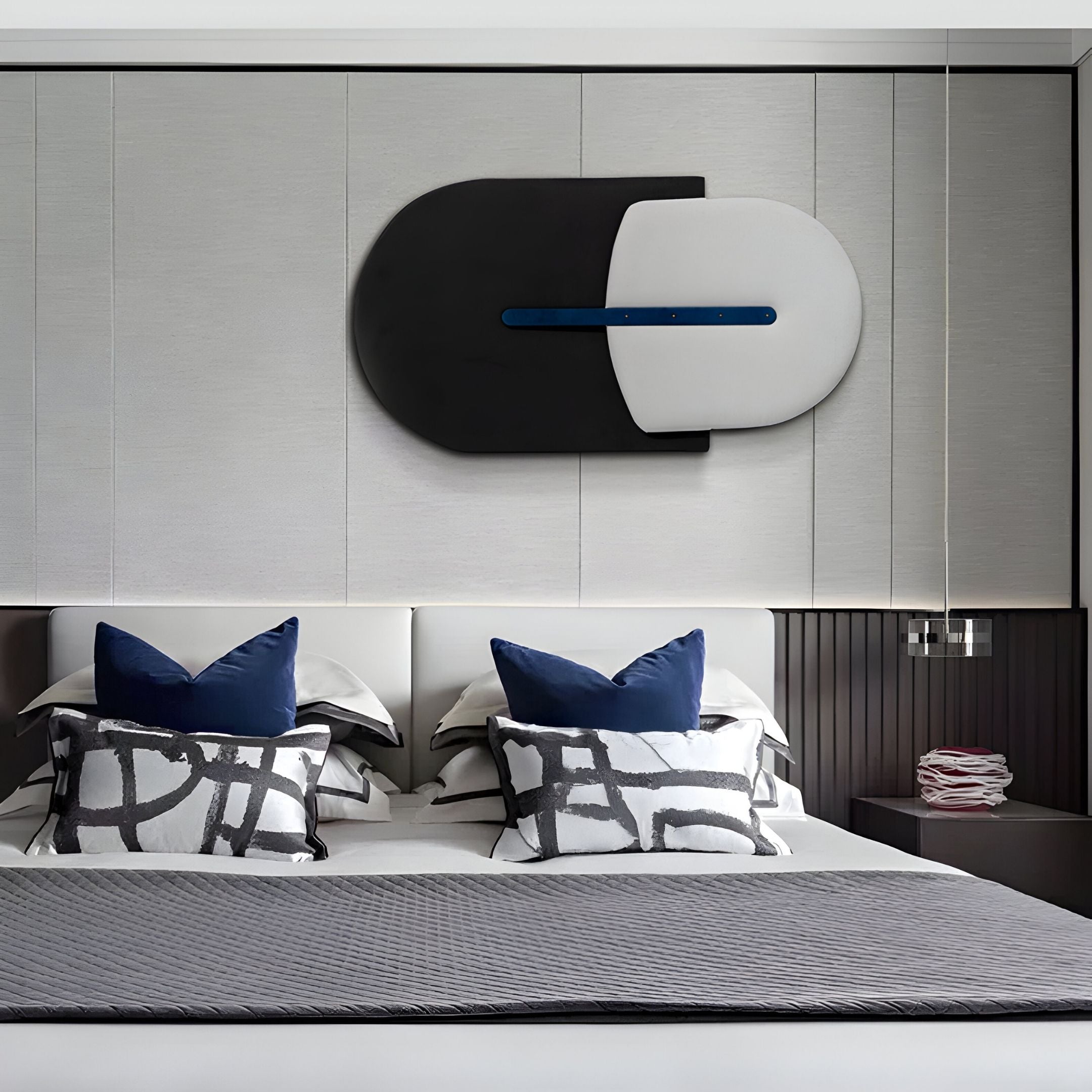 A modern bedroom features Solvia Black & White Layered Geometry 3D Wall Art by Giant Sculptures above the bed, with blue accent pillows, patterned black and white cushions, a gray quilt, and a stack of books on a bedside table near dark paneling.