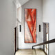 A modern hallway features the Rivara Ember Wave Vertical Hand-Painted Rope 3D Wall Art by Giant Sculptures in bold red and orange hues on a white wall, framed by black sconces as sunlight casts dramatic shadows across the space.