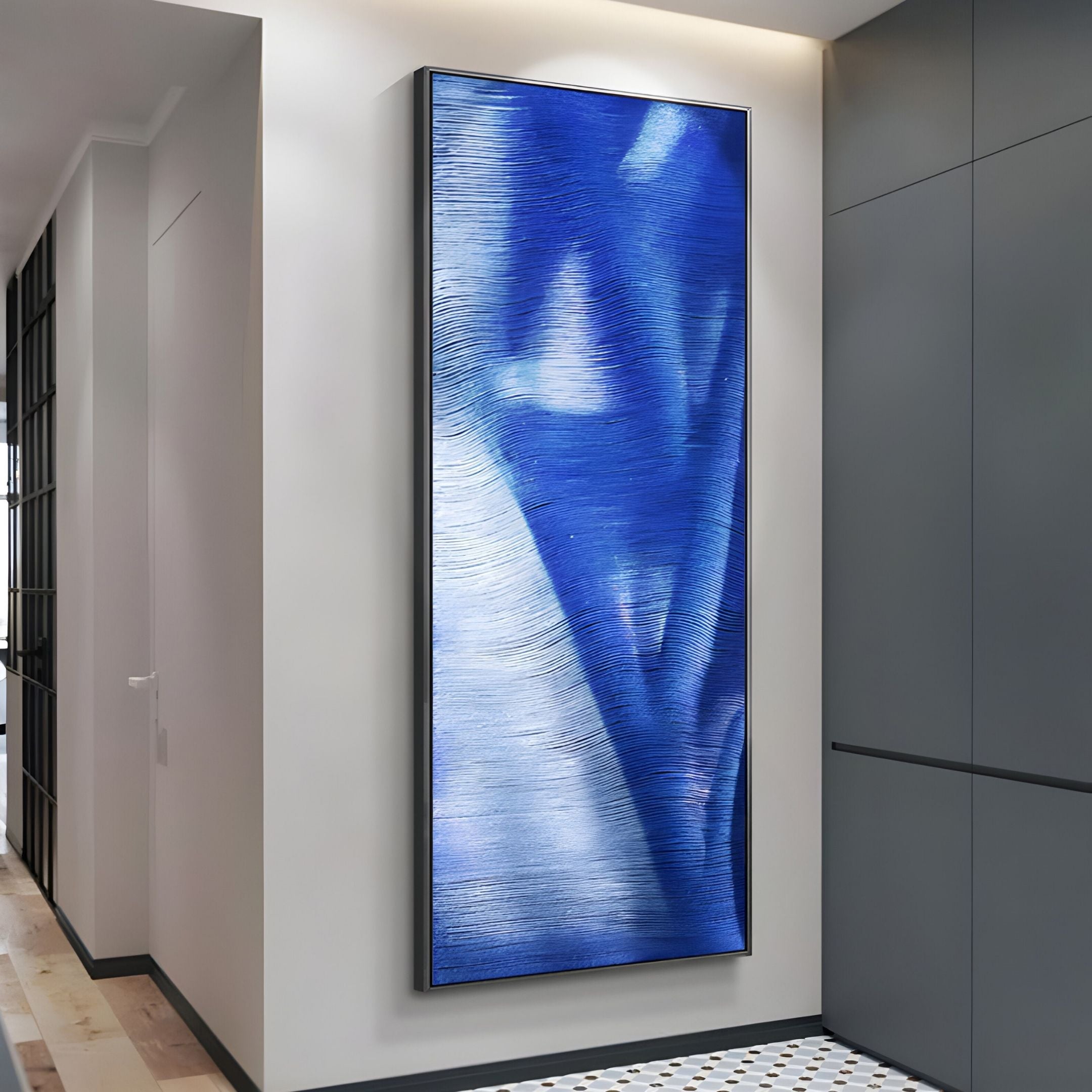 The Rivara Cobalt Wave Vertical Hand-Painted Rope 3D Wall Art by Giant Sculptures, in blue and white, hangs in a modern hallway with neutral decor and geometric tiled flooring.