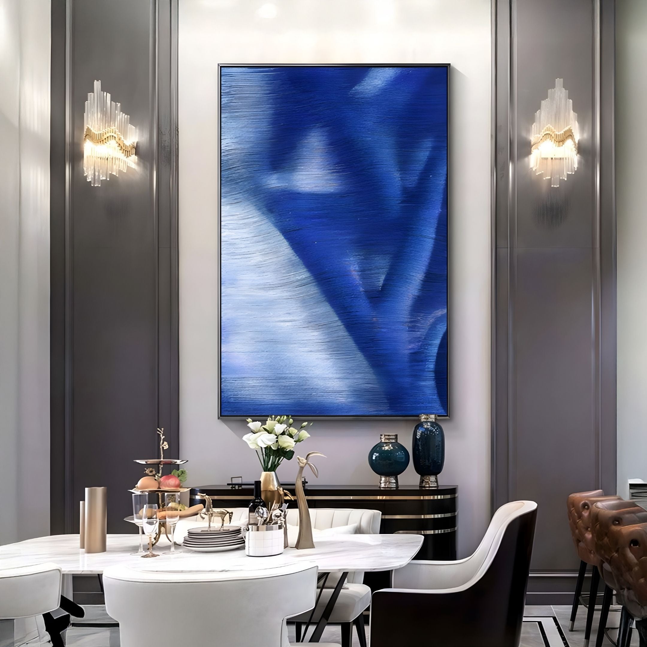 A modern dining room showcases Giant Sculptures' Rivara Cobalt Flow Hand-Painted Rope 3D Wall Art, elegant wall sconces, a round marble table with white chairs, plus decorative vases and flowers on a sideboard.
