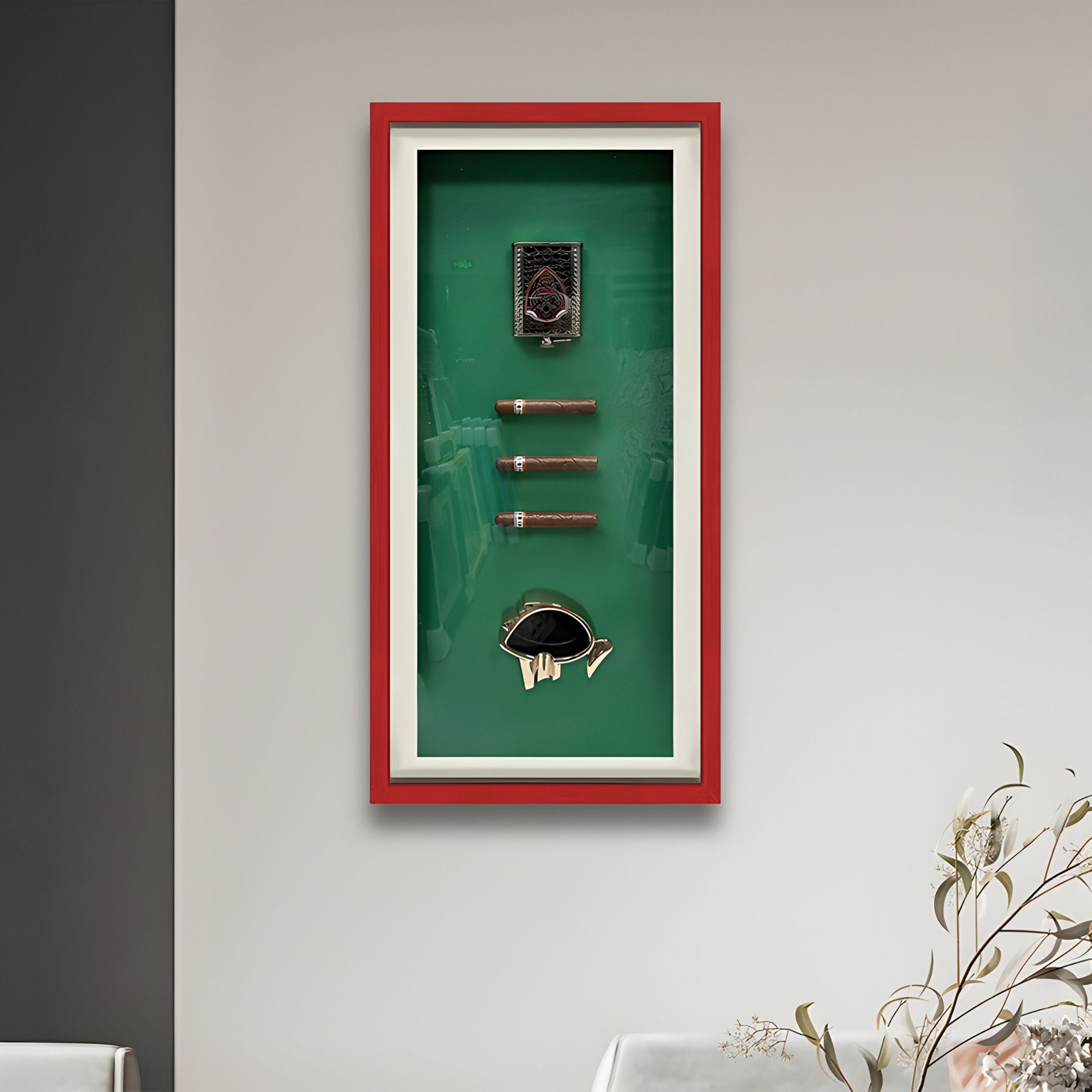 The Cavendor Cigar & Badge Green Framed Wall Art by Giant Sculptures features three cigars, a lighter, cutter, and ashtray on a green background with a red border. A plant sits in the lower right corner against a gray wall.