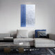 Modern living room with a gray sectional, glass coffee table, and Giant Sculptures’ Metallure Blue Linear Textured Acrylic Silver Foil 3D Wall Art. Decorative pillows, books, and a dark patterned rug complete the minimalist decor.