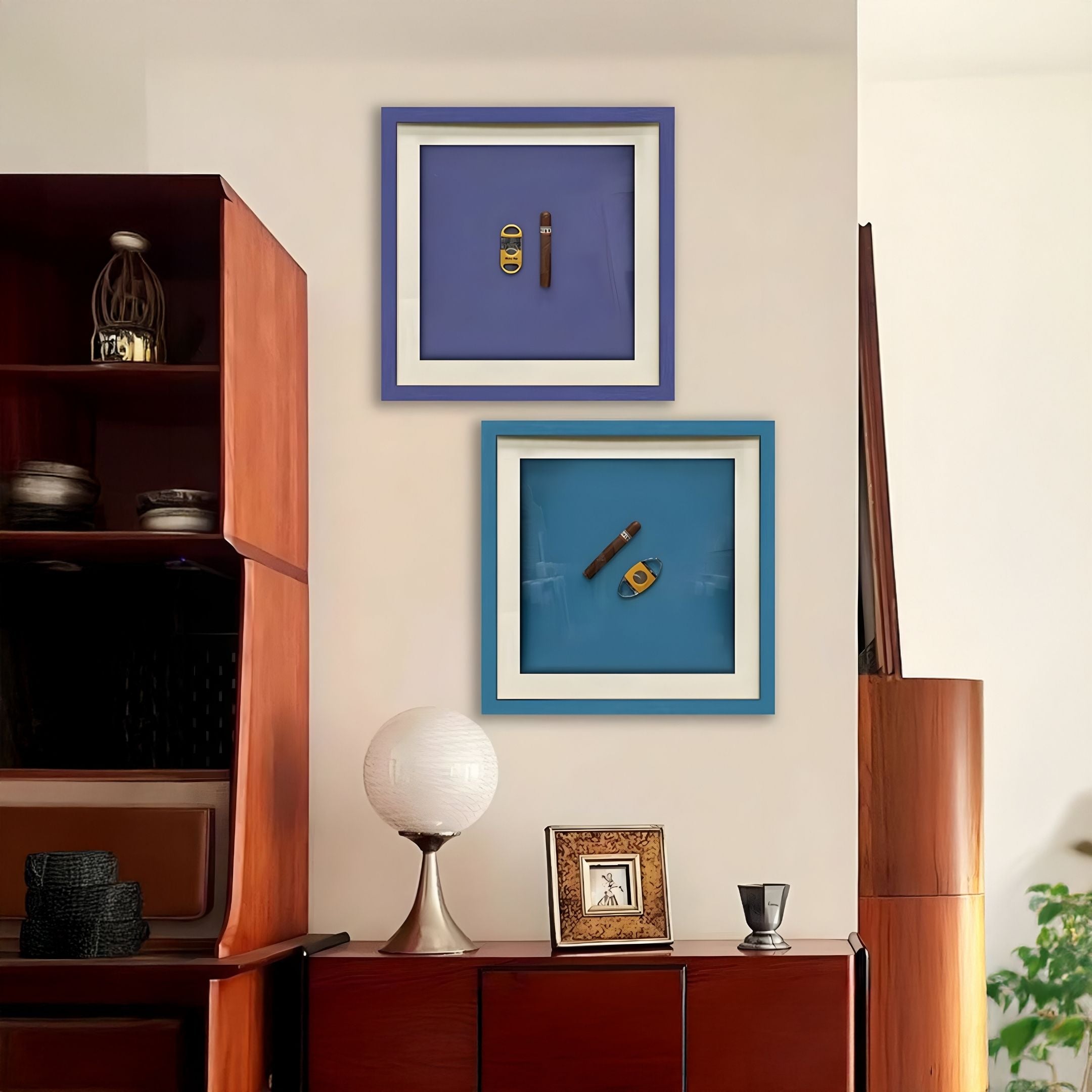 A modern living space features wooden shelves, decorative items, a globe lamp, and two Cavallero Blue Cigar & Cutter Framed Wall Art pieces by Giant Sculptures, each showcasing unique objects on blue backgrounds as contemporary displays.