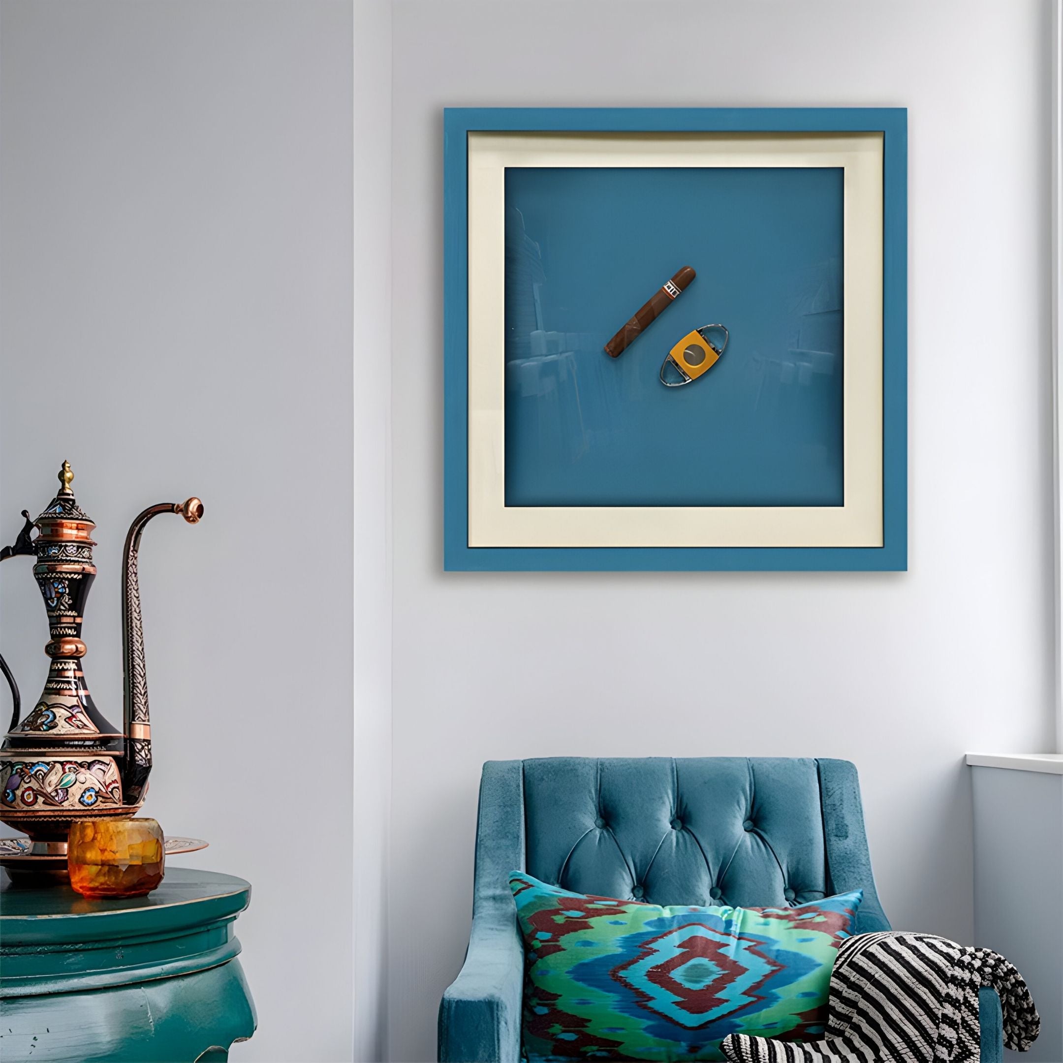 A cozy corner with a blue velvet chair and colorful pillow, beside an ornate metal teapot on a round green table. Above, Giant Sculptures' Cavallero Teal Cigar & Cutter Framed Wall Art brings a stylish modern decor touch.