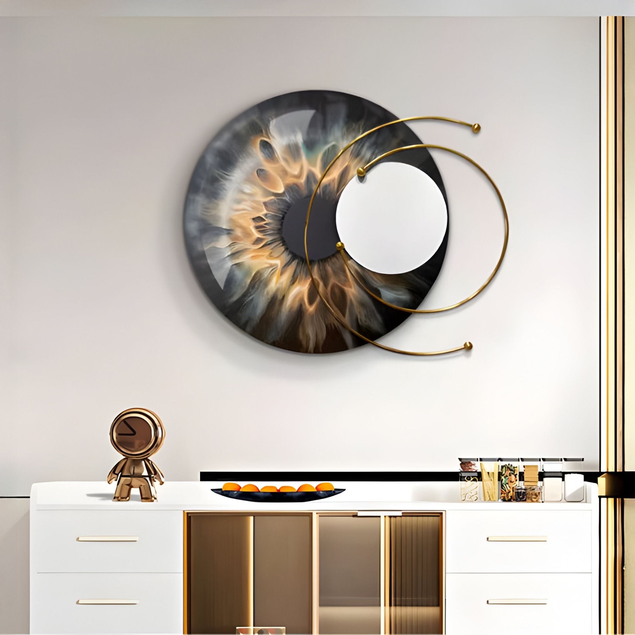 A modern interior with a white cabinet, gold accents, an abstract statue, a bowl of oranges, and the striking Aurelios Amber Flare Crystal Porcelain & Gold Orbit 3D Wall Art by Giant Sculptures—its large circular form showcases metallic and smoky hues with gold lines.