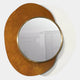 The Solistra Amber II Layered Round Mirror 3D Wall Art by Giant Sculptures features a gold-framed round mirror on abstract backplates in rusty orange and white, reflecting a soft image of a cabinet and books in a bright room.