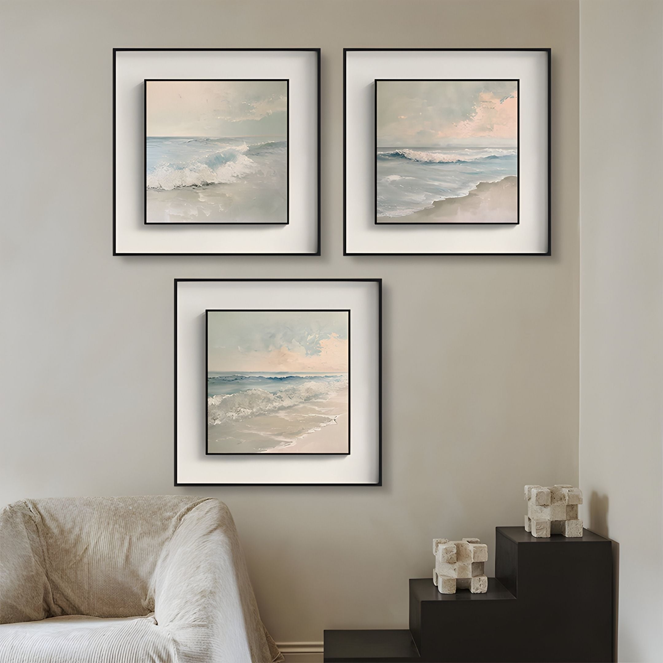 A cozy living room corner features a covered armchair, black stairs with decor, and three Giant Sculptures Tidalume IV Soft Dawn Shoreline Double Framed Wall Art pieces, showcasing serene ocean scenes on a light beige wall.