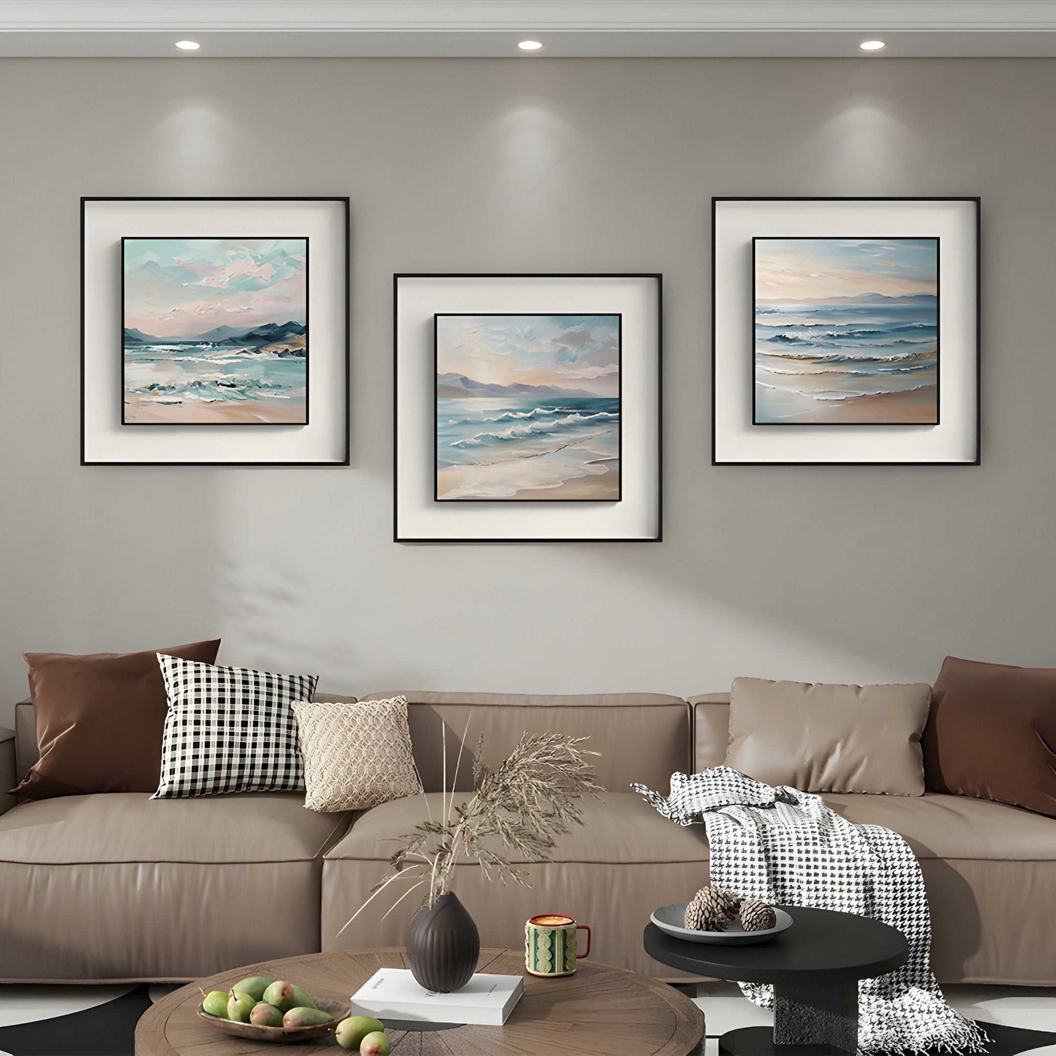 A modern living room with a beige couch, brown and checked pillows, a throw blanket, a coffee table with a vase and fruit, and three Marilline I Calm Coastal View Double Framed Wall Art pieces by Giant Sculptures on a light gray wall.