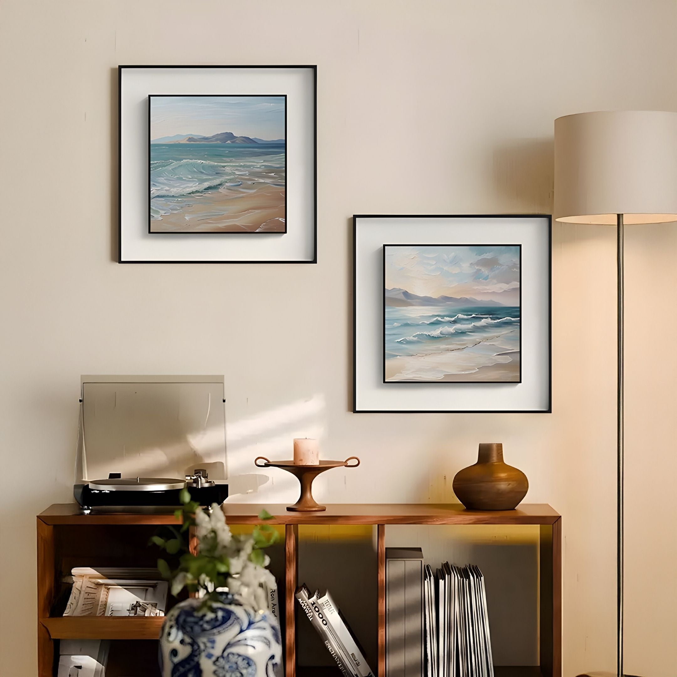 A cozy living room features the Giant Sculptures Marilline IV Calm Coastal View Double Framed Wall Art on a beige wall, above a wooden shelf displaying a record player, vases, candle, lamp, and neatly stacked magazines below.