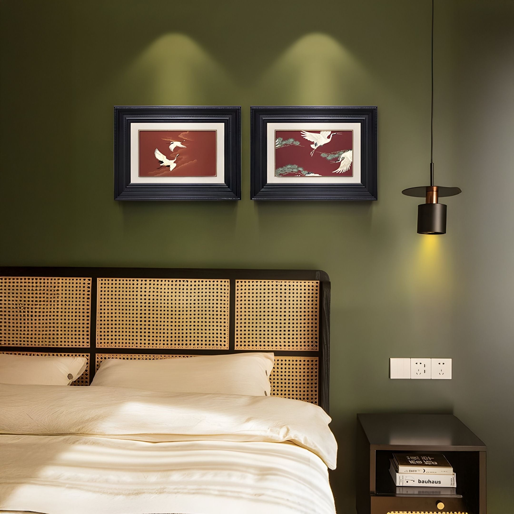 A bed with a wicker headboard sits against a green wall. Above it hangs the Kansora Heritage Crane Flight 2-Set Framed Wall Art by Giant Sculptures, while a black pendant lamp illuminates the modern bedside table.