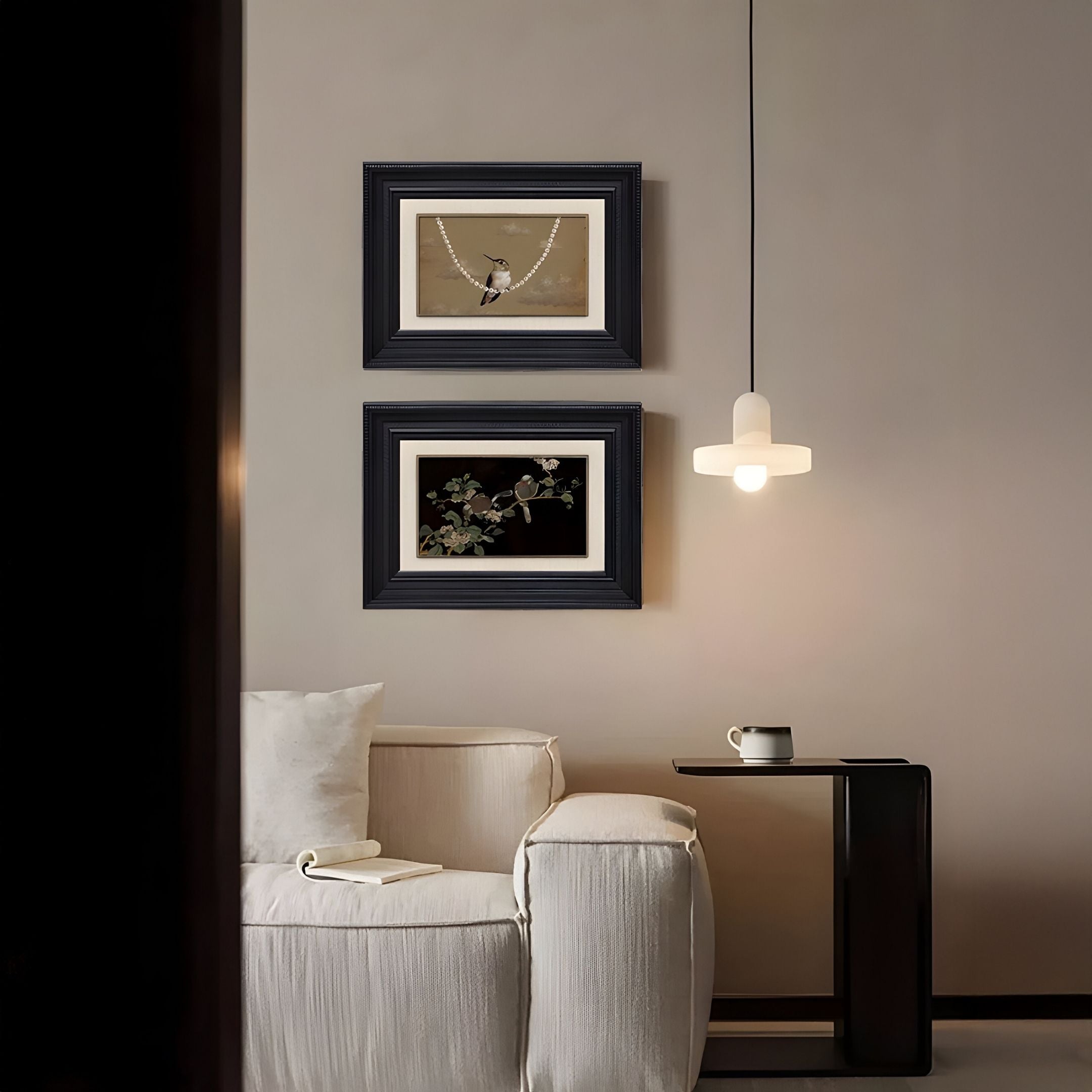A modern, minimal living room with a white sofa, a small black side table with a mug, and Giant Sculptures’ Kansora Songbird Pearl & Branch 2-Set Framed Wall Art featuring birds and botanicals above, plus a softly glowing pendant light.