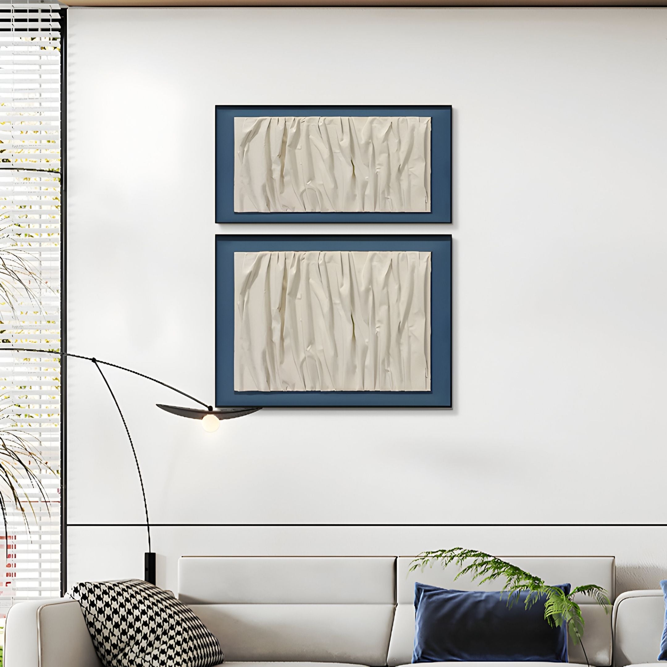 A modern living room with a white sofa, patterned pillow, and navy cushion. Above the sofa hangs the Foldara Cream & Blue Draped Paper Dual-Panel 3D Wall Art by Giant Sculptures. A floor lamp and green plant complete the setting.