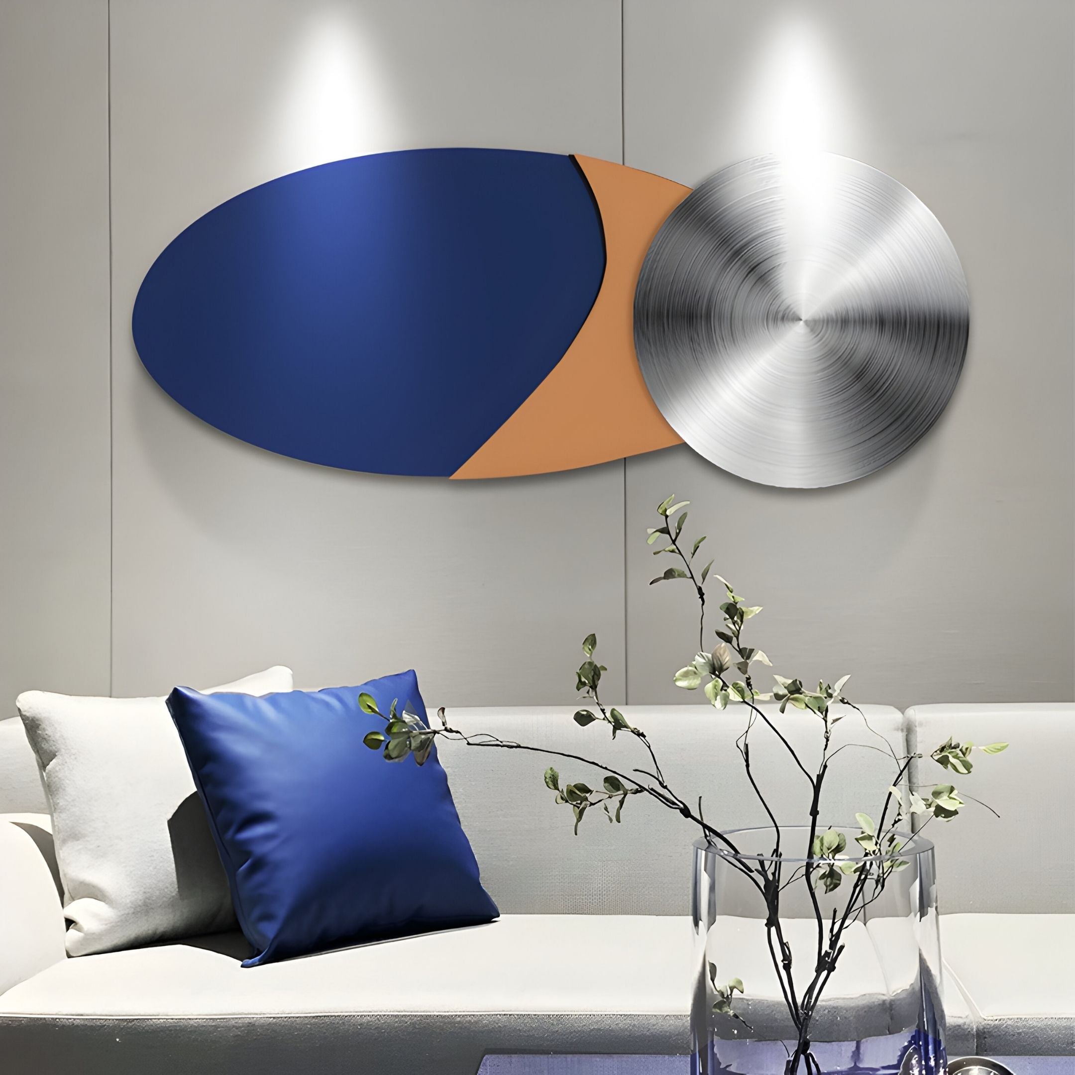A modern living room with a light sofa, blue pillow, glass vase with green branches, and the Aurelune Midnight Blue & Tan Silver Metal Disc 3D Wall Art by Giant Sculptures—striking decor for contemporary interiors.