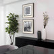 A modern minimalist interior features a black sideboard, decorative vases, a tall leafy plant, and two Stratonis Monochrome Abstract Textured Framed Wall Art pieces by Giant Sculptures on a white wall, with a gray chair and table visible in the foreground.