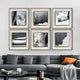 A modern living room with a gray sectional sofa, orange and black cushions, a round coffee table, and six Stratonis Grey Terrain Textured Framed Wall Art pieces by Giant Sculptures arranged above the sofa.
