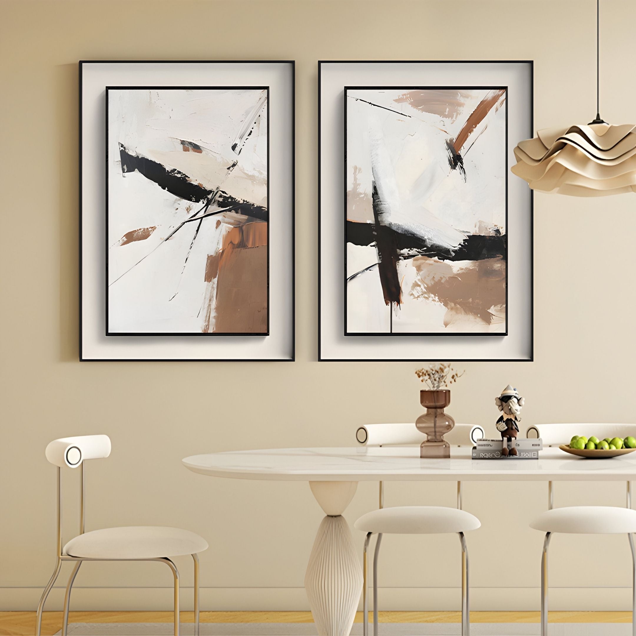 A modern dining area with a white oval table, white chairs, a bowl of green apples, and Giant Sculptures’ Bravura II Stroke Abstract Framed Canvas Wall Art—two bold black, brown, and white paintings—on a beige wall. A sculptural pendant light hangs above.