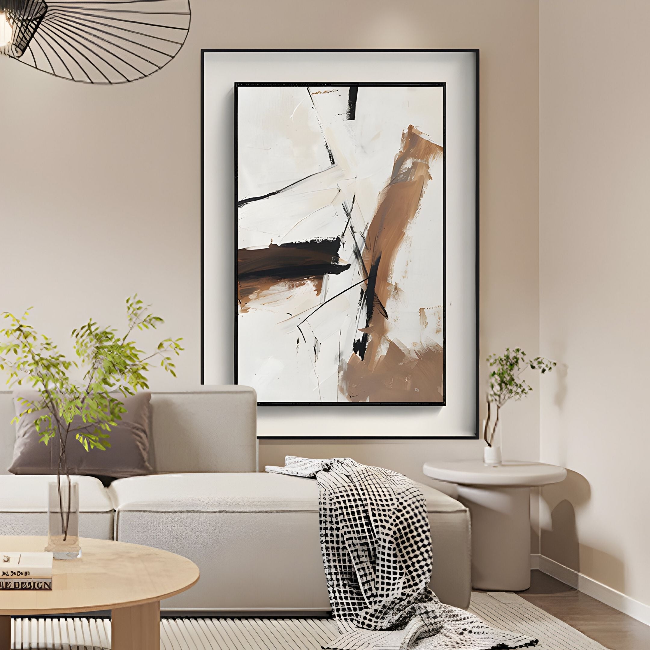 A modern living room with a beige couch, plaid blanket, potted plants, and round coffee table is accented by the Bravura III Stroke Abstract Framed Canvas Wall Art by Giant Sculptures in black, brown, and white tones above the sofa for a stylish touch.